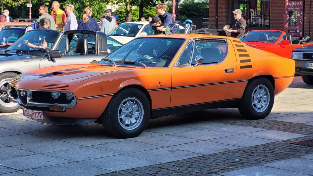 Ein orangefarbener Oldtimer-Sportwagen ist auf einer Autoshow im Freien geparkt. Im Hintergrund sind mehrere Personen und andere Autos zu sehen.